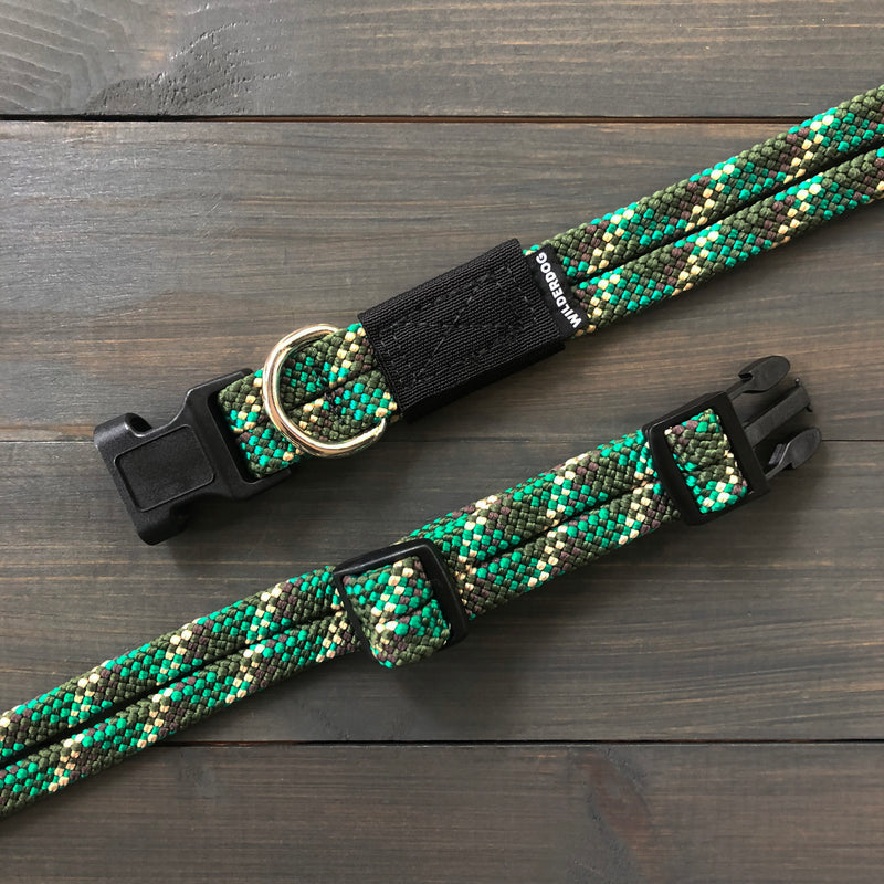 A close-up of two Camo Collars in green and black, featuring woven heavy-duty material with plastic buckle closures and metal D-rings, laid flat on a dark wooden surface. These collars have a textured pattern reminiscent of rock climbing rope and adjustable lengths with sliding buckles.